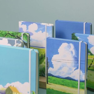 May include: A collection of colorful notebooks with landscape scenes on the covers. The notebooks have elastic bands and are stacked on a wooden surface.