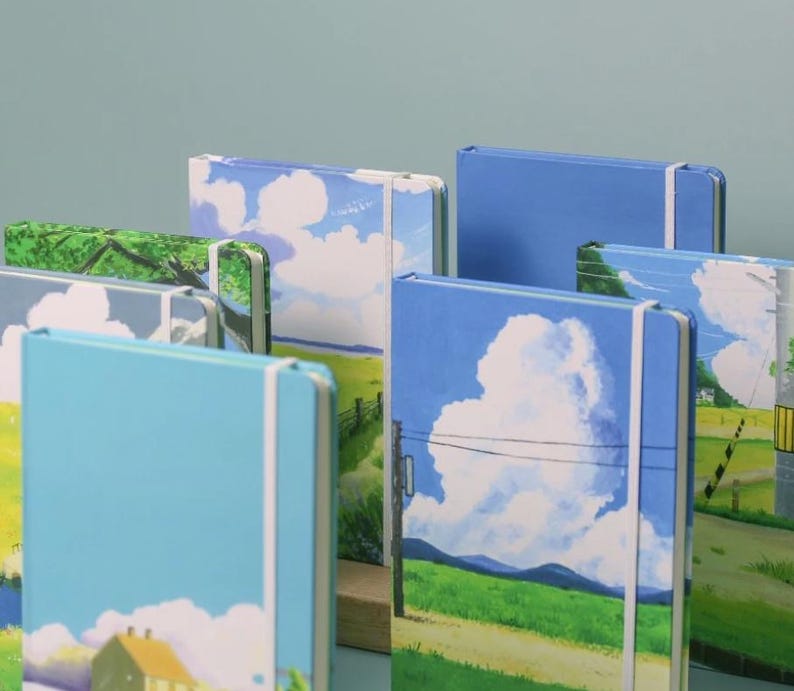 May include: A collection of colorful notebooks with landscape scenes on the covers. The notebooks have elastic bands and are stacked on a wooden surface.