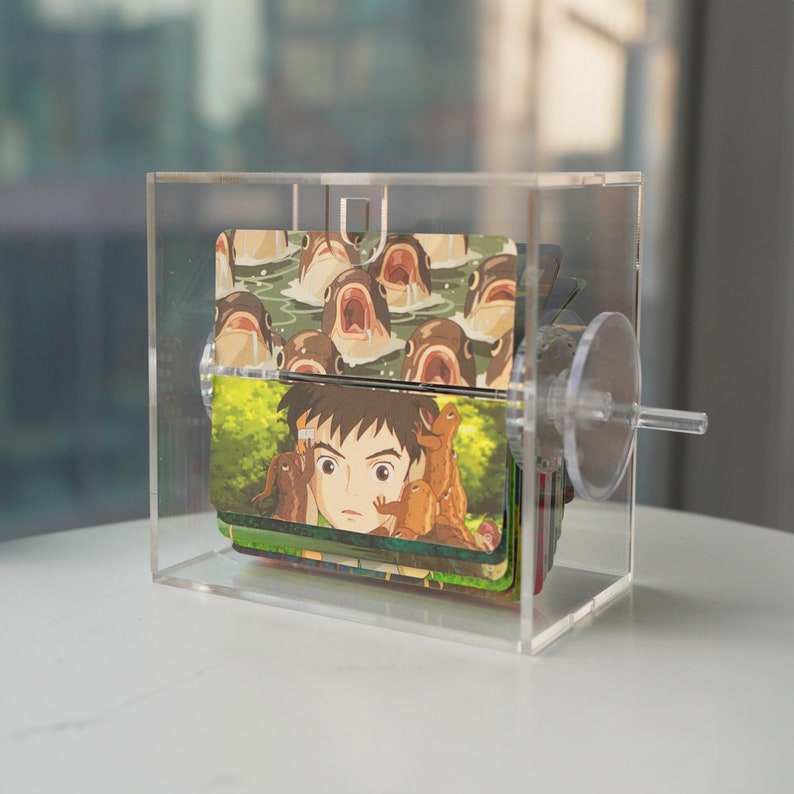 K&ouml;nnte beinhalten: Eine transparente Acrylbox mit einem Drehmechanismus enth&auml;lt einen Stapel bunter Karten mit Comicbildern von Fischen und einem Jungen. Die Karten sind &uuml;bereinander gelegt und durch die transparenten Acrylseiten der Box sichtbar.