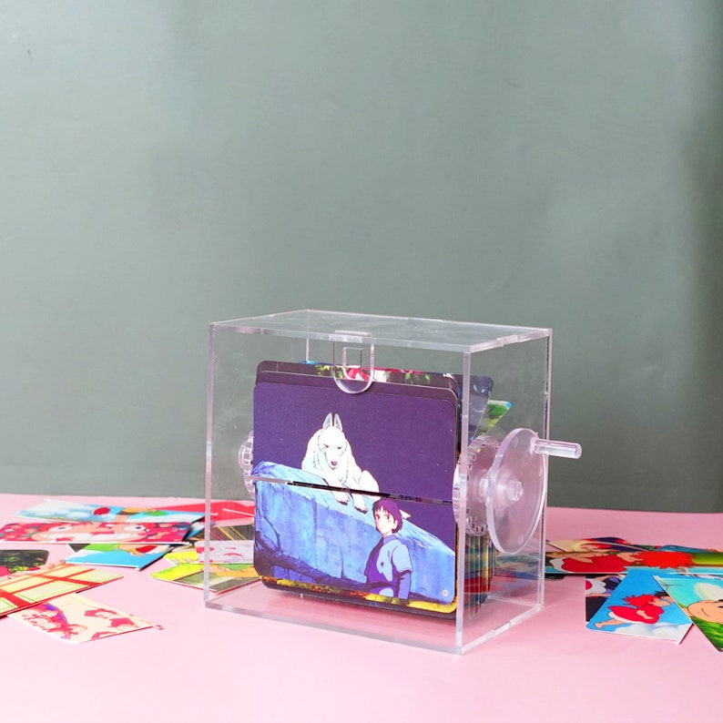 Puede incluir: Una caja de acr&iacute;lico transparente con una manivela en el lateral, que contiene una pila de tarjetas coloridas con im&aacute;genes de la pel&iacute;cula animada "Mi vecino Totoro".