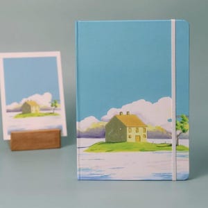 May include: A blue notebook with a white elastic band closure features a hand-painted illustration of a yellow house on a green island with a blue sky and white clouds.