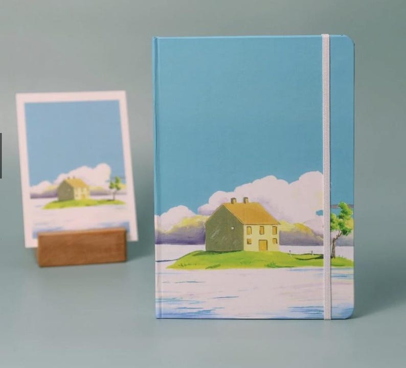May include: A blue notebook with a white elastic band closure features a hand-painted illustration of a yellow house on a green island with a blue sky and white clouds.