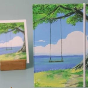 May include: A green, blue, and white painted notebook with a swing hanging from a tree branch over a blue ocean. The notebook has a white elastic closure and a white ribbon bookmark.