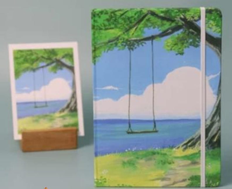May include: A green, blue, and white painted notebook with a swing hanging from a tree branch over a blue ocean. The notebook has a white elastic closure and a white ribbon bookmark.