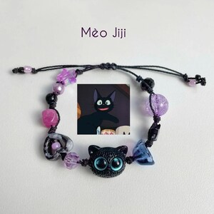 Puede incluir: Un brazalete negro y morado con un dije de gato negro y una peque&ntilde;a imagen de un gato negro con ojos grandes. El brazalete tiene una variedad de cuentas, incluyendo estrellas, corazones y cuentas redondas.