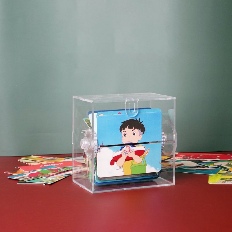 May include: A clear acrylic box with a rotating display of colorful cards featuring cartoon characters. The cards are stacked and visible through the clear sides of the box.