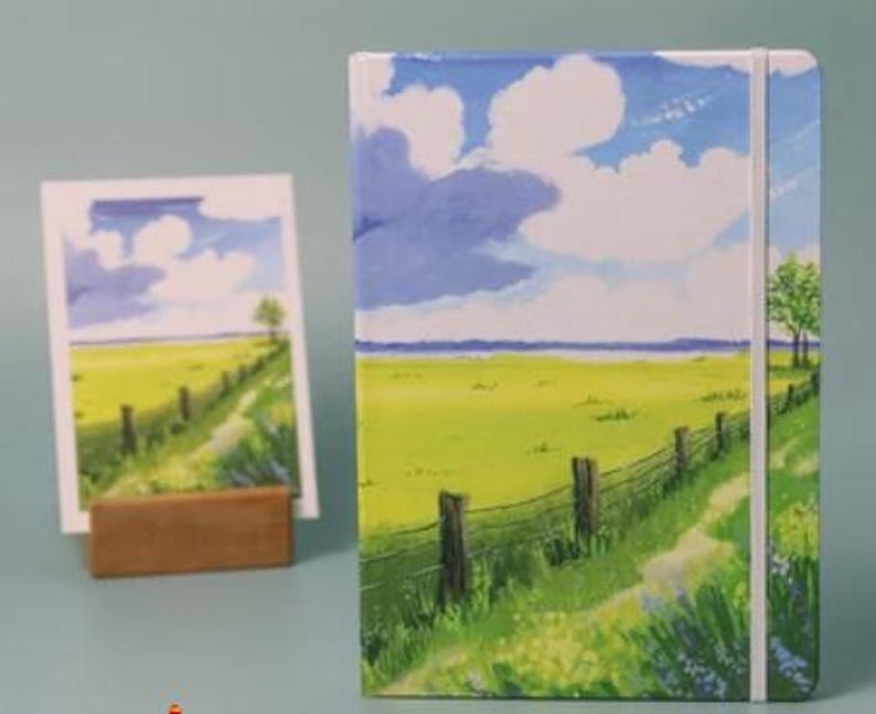May include: A landscape notebook with a white cover and a blue sky with white clouds. The cover features a painting of a green field with a wooden fence and a path leading to a distant horizon.