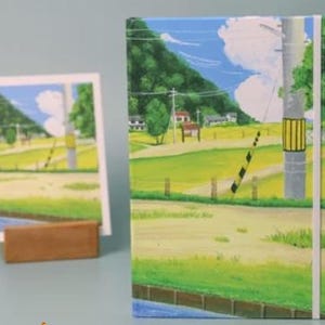 May include: A colorful notebook with a landscape scene of a rural road, a wooden fence, and a power line. The scene is painted in a cartoon style with bright green grass, yellow fields, and a blue sky with white clouds.