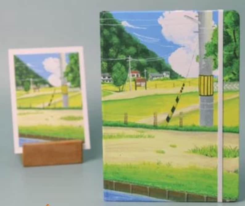 May include: A colorful notebook with a landscape scene of a rural road, a wooden fence, and a power line. The scene is painted in a cartoon style with bright green grass, yellow fields, and a blue sky with white clouds.