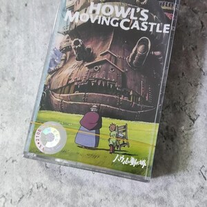 Pu&ograve; includere: Un nastro audio vintage con un'immagine colorata di un grande castello mobile dal film "Il castello errante di Howl". Il castello &egrave; marrone e grigio con una grande bocca aperta. Due figure, una con un vestito verde e l'altra con un vestito marrone, si trovano davanti al castello. Il testo "Il castello errante di Howl" &egrave; stampato in bianco su sfondo nero. Il testo "八九〇番" &egrave; stampato in nero su sfondo bianco.