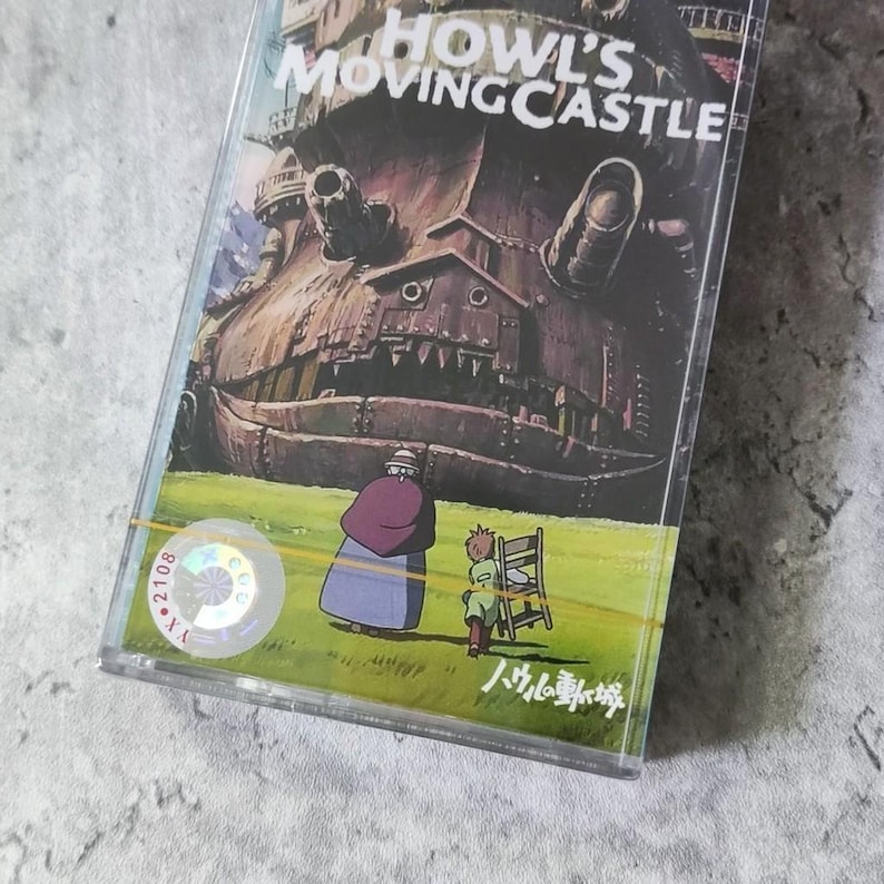 Pu&ograve; includere: Un nastro audio vintage con un'immagine colorata di un grande castello mobile dal film "Il castello errante di Howl". Il castello &egrave; marrone e grigio con una grande bocca aperta. Due figure, una con un vestito verde e l'altra con un vestito marrone, si trovano davanti al castello. Il testo "Il castello errante di Howl" &egrave; stampato in bianco su sfondo nero. Il testo "八九〇番" &egrave; stampato in nero su sfondo bianco.
