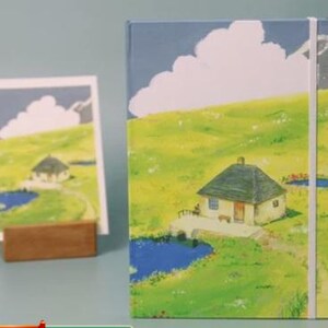 May include: A blue and green illustrated notebook with a white cloud and a small house with a brown roof. The notebook has a white elastic band closure.