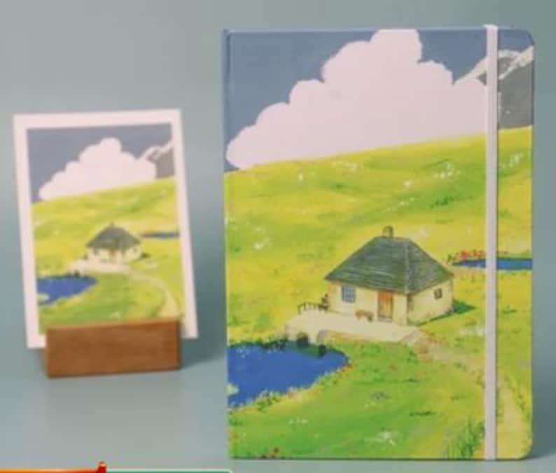 May include: A blue and green illustrated notebook with a white cloud and a small house with a brown roof. The notebook has a white elastic band closure.