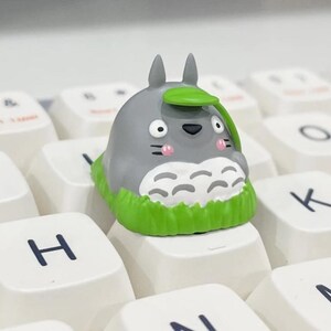 May include: A custom keycap featuring a gray Totoro character with a green leaf hat and grass base, placed on a white keyboard. The keycap has a cartoon design with pink cheeks and is surrounded by white keys with black lettering.