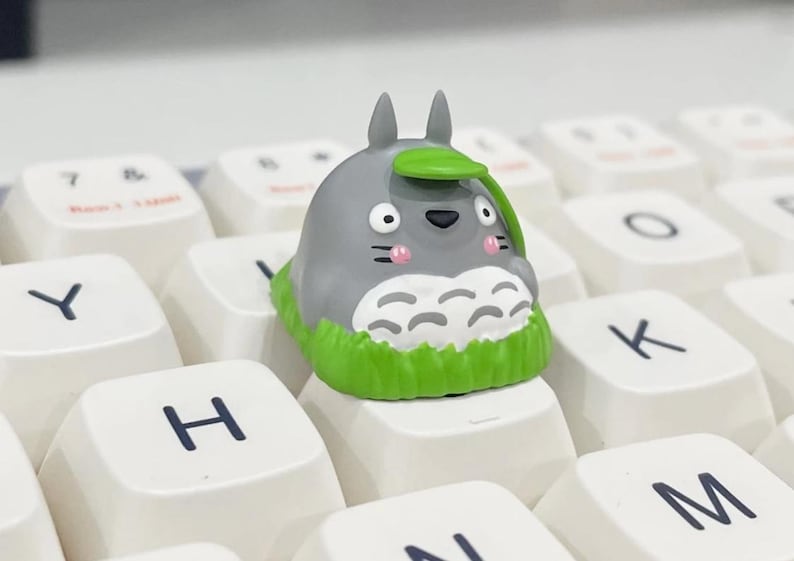 May include: A custom keycap featuring a gray Totoro character with a green leaf hat and grass base, placed on a white keyboard. The keycap has a cartoon design with pink cheeks and is surrounded by white keys with black lettering.