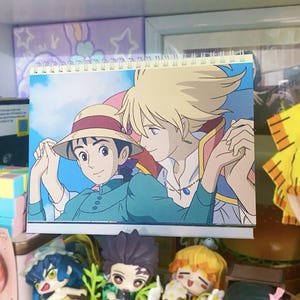 Może przedstawiać: Spiralnie oprawiony kalendarz z ilustracją w stylu anime dw&oacute;ch postaci. Jedna postać nosi kapelusz i zielone ubrania, druga ma blond włosy i białą koszulę. Tło to błękitne niebo z chmurami.
