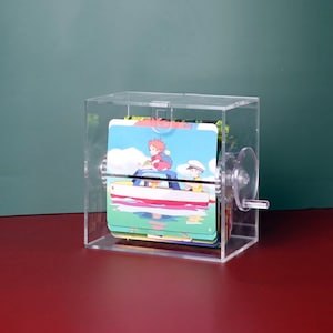 May include: A clear acrylic box with a crank on the side. Inside the box are several cards with illustrations of a girl and boy in a boat. The cards are stacked on top of each other.