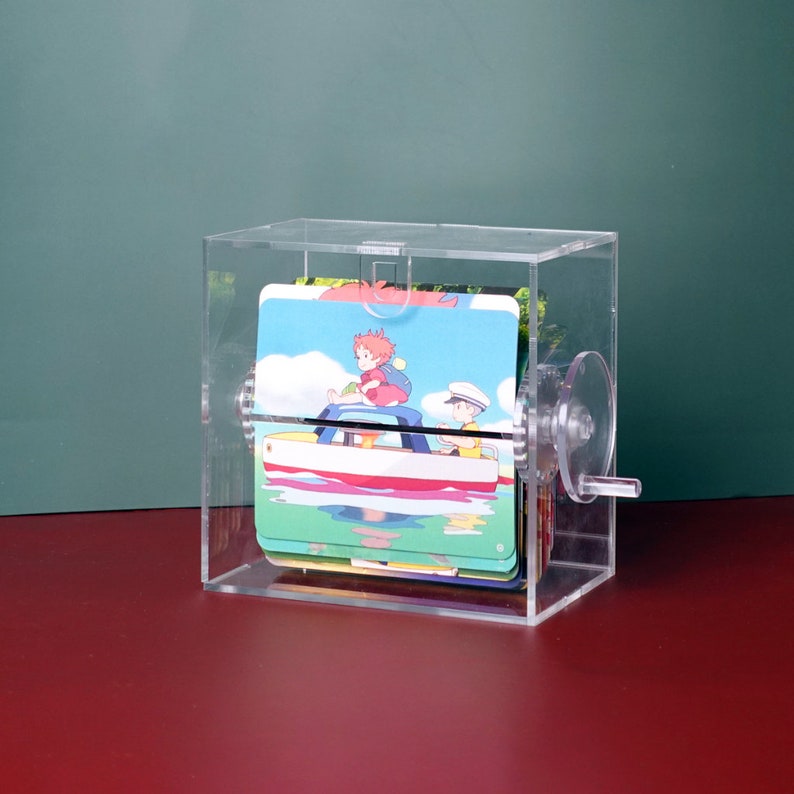 May include: A clear acrylic box with a crank on the side. Inside the box are several cards with illustrations of a girl and boy in a boat. The cards are stacked on top of each other.