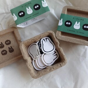 May include: A set of 14 paper clips in two designs, featuring a white and black cartoon character. The clips are packaged in a small, brown cardboard box with a green label that reads "P-Clips 2 Designs 14 Pieces".