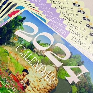 May include: A 2024 calendar featuring a watercolor illustration of a green meadow with a thatched roof hut. The calendar is decorated with a butterfly and the text "COSY GHIBLI" in a whimsical font. The calendar is divided into 12 months, with each month labeled in Vietnamese.