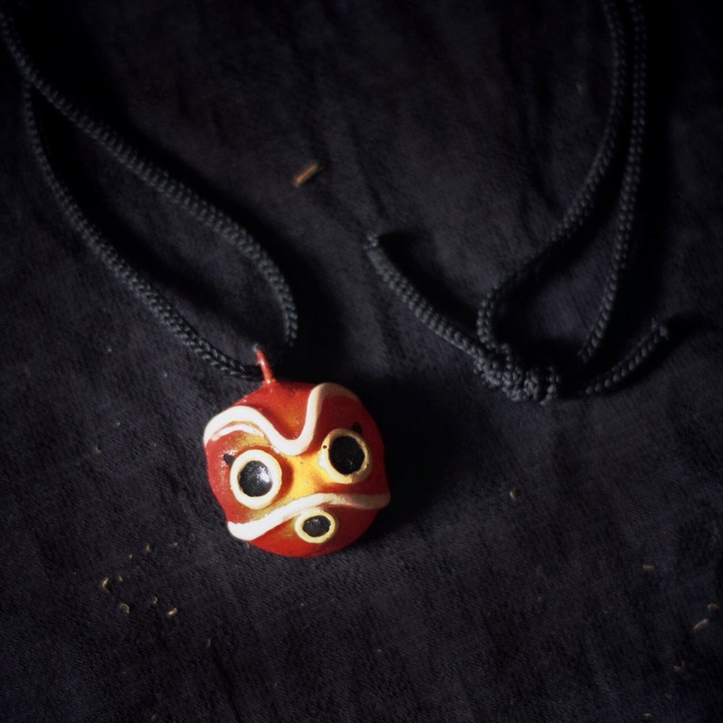 May include: A red and white mask with black eyes and a white mouth hangs on a black cord. The mask is a popular character from the animated film Spirited Away.