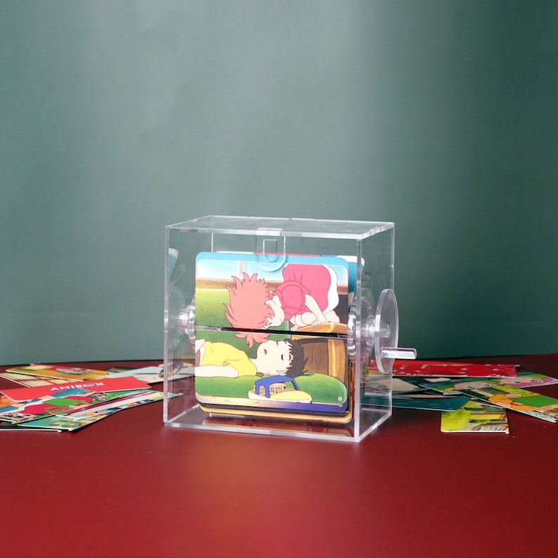 May include: A clear acrylic box with a rotating mechanism, displaying a card with an illustration of two cartoon characters. The card is part of a set of cards that can be rotated through the box.