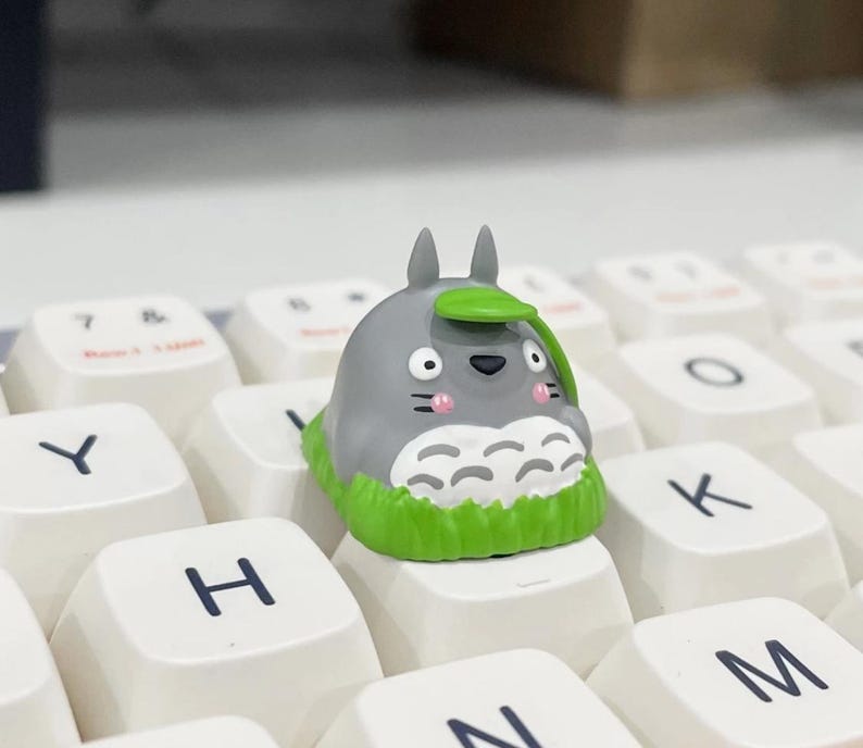 May include: A gray Totoro-shaped keycap with a green leaf on its head, sitting on a white keyboard key. The keycap has a green grass-like base and pink cheeks. The keyboard keys are white with black lettering.