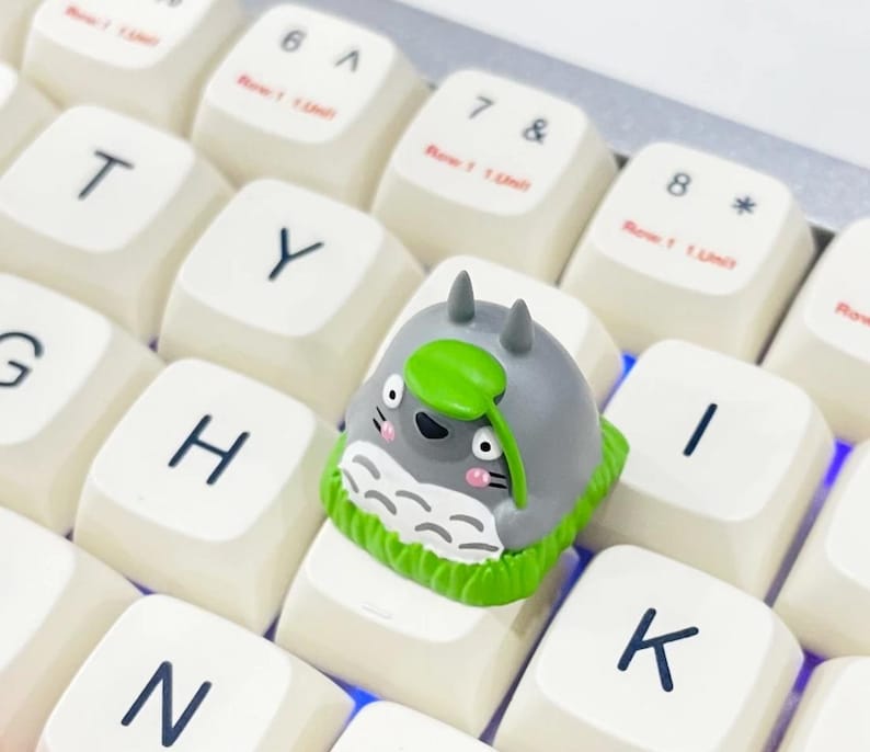 May include: A custom keycap featuring a gray Totoro character with a green leaf, set on a white keyboard. The keycap is surrounded by other white keys with black lettering. The keyboard is lit with a blue glow.