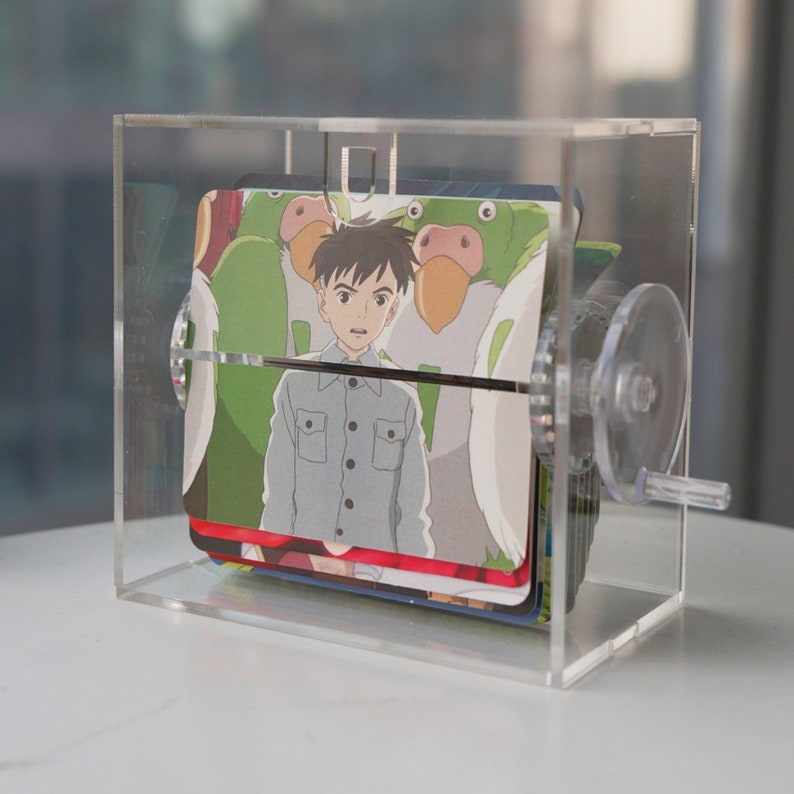 K&ouml;nnte beinhalten: Eine transparente Acrylbox mit einer Kurbel an der Seite, die einen Stapel bunter Karten mit Anime-Illustrationen enth&auml;lt. Die oberste Karte zeigt einen jungen Jungen mit braunen Haaren und einem gr&uuml;n-blau-rot gemusterten Hemd, der vor einer Gruppe von Comicfiguren steht.