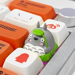 May include: A custom keyboard with orange, white, and gray keycaps. A gray Totoro keycap with a green leaf sits among the other keys. The keyboard also has a silver volume knob.