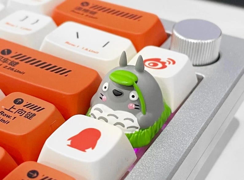 May include: A custom keyboard with orange, white, and gray keycaps. A gray Totoro keycap with a green leaf sits among the other keys. The keyboard also has a silver volume knob.