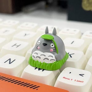 May include: A gray Totoro-themed keycap with a green leaf and grass detail, sitting on a white keyboard. The keycap has pink cheeks and a black nose and eyes. The keyboard keys are white.