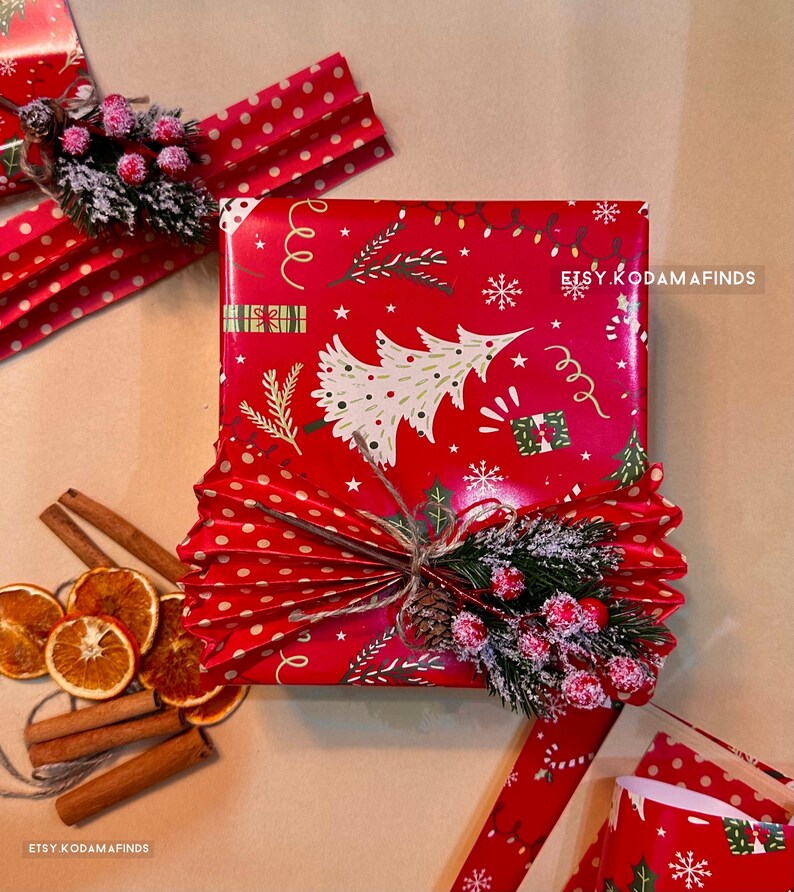 K&ouml;nnte beinhalten: Eine rote Geschenkbox, verpackt in Weihnachtspapier mit einem wei&szlig;en und gr&uuml;nen Weihnachtsbaumdesign. Die Box ist mit einem braunen Bindfaden gebunden und mit einem gr&uuml;nen Tannenzweig mit roten Beeren und einem wei&szlig;en Papierf&auml;cher verziert.