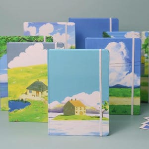 May include: A set of six notebooks with different landscape illustrations. Each notebook has a white elastic band closure and a blue, green, or white cover. The covers feature illustrations of houses, fields, and clouds.