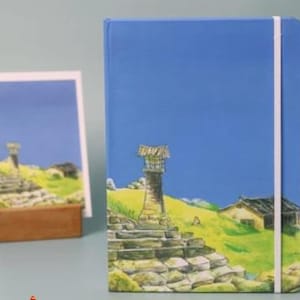 May include: A blue notebook with a white elastic closure and a colorful illustration of a stone tower, a small house, and a blue sky.