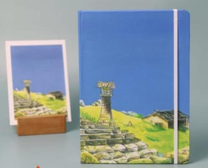 May include: A blue notebook with a white elastic closure and a colorful illustration of a stone tower, a small house, and a blue sky.