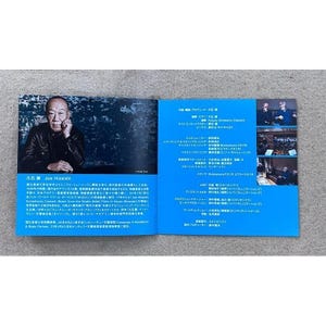 May include: A blue and white booklet with a photo of a man on the left side and text on the right side. The text includes the name "Joe Hisaishi" and the title "Future Orchestra Classics".