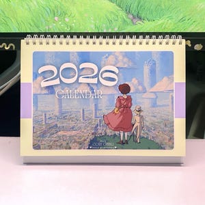 Może przedstawiać: Kalendarz na rok 2026 z fantastyczną ilustracją. Okładka przedstawia scenę z dwiema postaciami spoglądającymi na miasto z wysokimi, grzybowatymi strukturami. Kalendarz ma spiralną oprawę i kremową podstawę.
