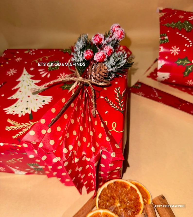May include: A red and white polka dot gift box with a red and green Christmas tree pattern. The box is tied with twine and decorated with a sprig of faux greenery, red berries, and a pine cone. The text "ETSY KODAMAFINDS" is printed on the box.
