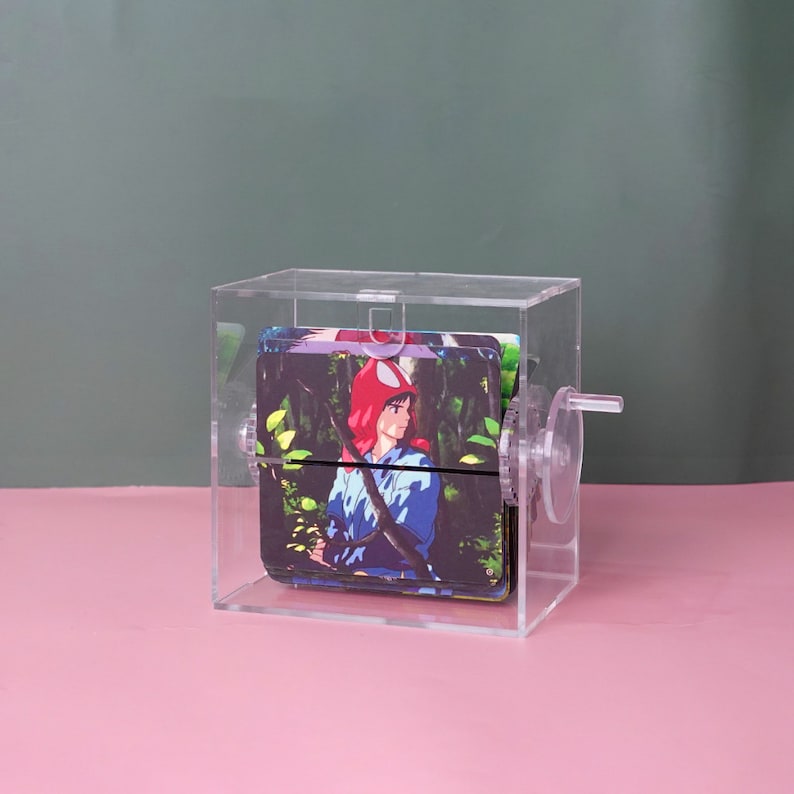 Puede incluir: Una caja de acr&iacute;lico transparente con un mecanismo giratorio, que muestra una pila de tarjetas coloridas con un personaje de anime de cabello rojo. El personaje lleva un atuendo azul y blanco y sostiene una espada.