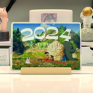 May include: A 2024 calendar featuring characters from the Studio Ghibli film My Neighbor Totoro. The calendar has a green and brown background with a depiction of Totoro and two children sitting in a field of flowers. The text "COSY GHIBLI" and "World of Dreams" is printed on the bottom of the calendar.
