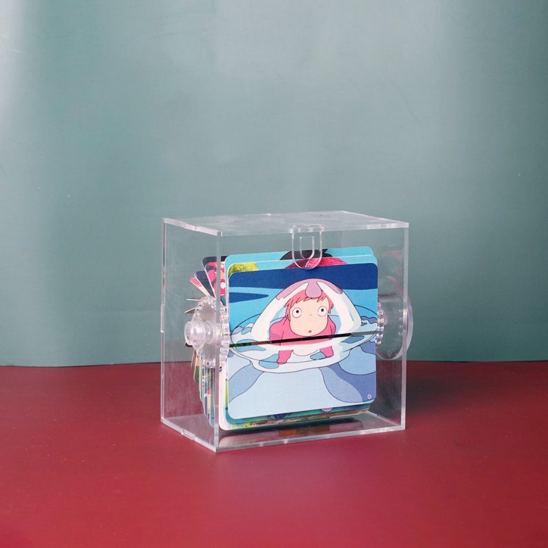May include: A clear acrylic box with a rotating mechanism, holding a stack of colorful cards featuring an anime character. The character is a pink creature with large eyes and a surprised expression, submerged in water.