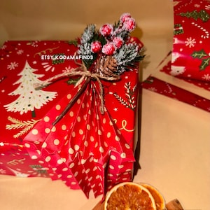 May include: A red and white polka dot gift box with a red and green Christmas tree design. The box is tied with twine and decorated with a sprig of faux greenery, red berries, and a pine cone. The text "ETSY KODAMAFINDS" is visible on the box.