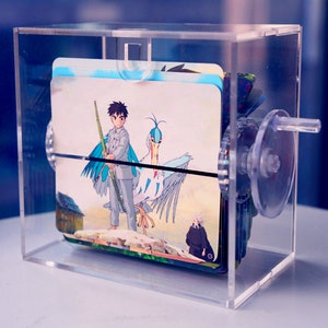 K&ouml;nnte beinhalten: Eine transparente Acrylbox mit einer Kurbel an der Seite. Im Inneren der Box befinden sich mehrere Karten mit Illustrationen eines jungen Menschen mit einem Vogel und einer Person in einem langen Mantel. Die Karten sind gestapelt und k&ouml;nnen mit der Kurbel gedreht werden.