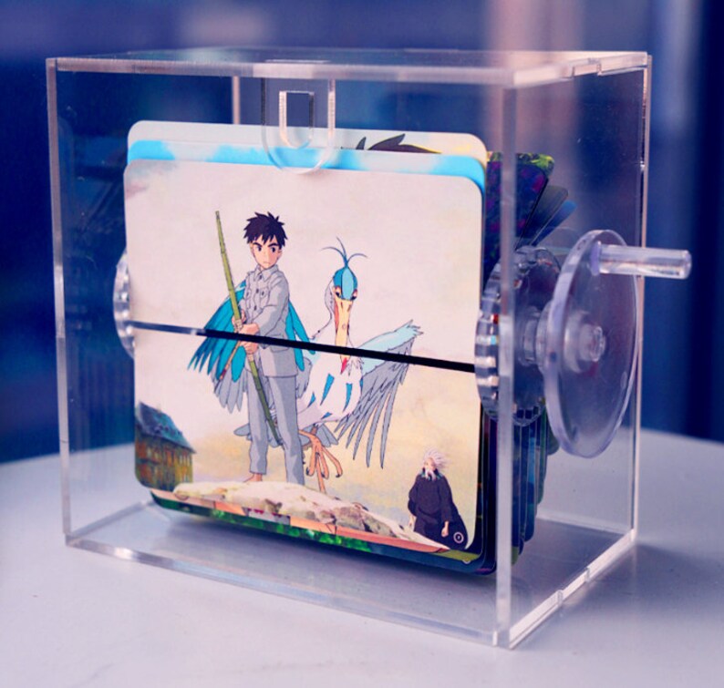 K&ouml;nnte beinhalten: Eine transparente Acrylbox mit einer Kurbel an der Seite. Im Inneren der Box befinden sich mehrere Karten mit Illustrationen eines jungen Menschen mit einem Vogel und einer Person in einem langen Mantel. Die Karten sind gestapelt und k&ouml;nnen mit der Kurbel gedreht werden.