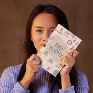 以下が含まれることがあります： DAILY PLANNERという暗い文字が中央の茶色の有機的な形の中に書かれた、ライトベージュの表紙のデイリープランナー。表紙には、植物、時計、カレンダー、本などの小さなカラフルなイラストが描かれています。