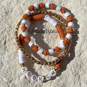 May include: A set of three bracelets with orange and white beads, and a gold chain with the word "HOOK 'EM" spelled out in white beads.