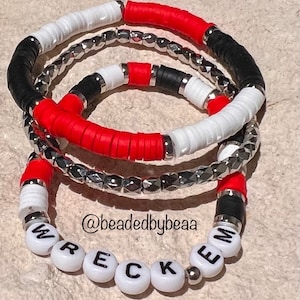 May include: Three layered bracelets with red, white, and black beads. The bottom layer is a silver chain with a silver bead between each link. The top layer spells out the word "WRECKEM" in white beads with black lettering.