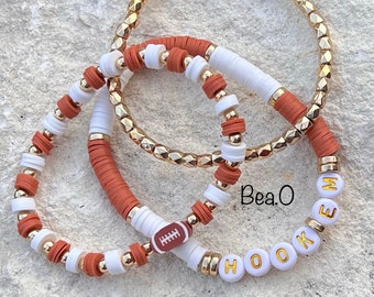 May include: Set of three bracelets with brown and white beads. One bracelet has a football charm, another has gold beads, and the third has the word "HOOK'EM" spelled out in white beads with gold lettering. The bracelets are on a light brown background.