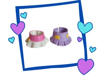Könnte beinhalten: Zwei Plastik-Puppenröcke, einer rosa mit weißem Besatz und einer lila mit gelben Akzenten.